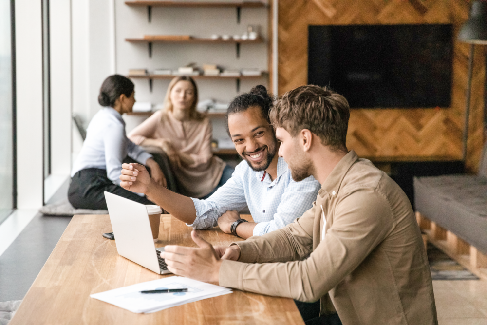 Zwei Männer, die an einem Tisch sitzen und sich unterhalten, auf dem Tisch liegt ein Blattpapier und ein Laptop.