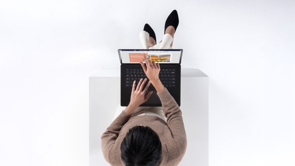 Eine Frau, die in einem weißen Raum auf einem weißen Quadrat sitzt. Sie hat einen Laptop auf ihrem Schoß und nutzt die Touch-Funktion ihres Displays.